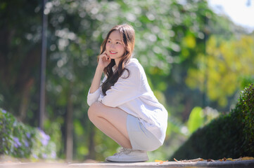A young beautiful Asian woman in a white shirt and light skirt crouches outdoors on a pathway. She is smiling and looking up with a blurred green park background.