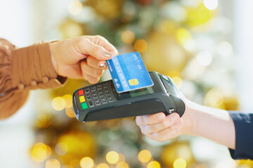 A close-up shows a hand with a blue credit card making a contactless payment over a card reader. The blurred background of warm lights suggests a festive or modern indoor retail transaction.