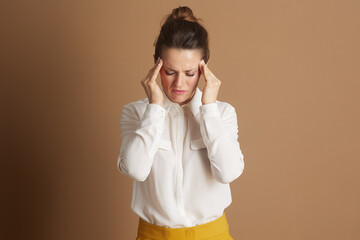 A professional woman in a white blouse and yellow skirt, eyes closed with hands pressed to her temples, depicts stress or a headache against a plain beige background.