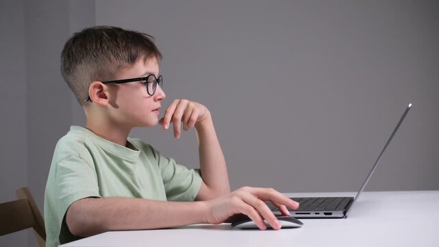 Young schoolboy in glasses intently using a laptop and mouse at a desk, engaged in remote learning and digital study, focused on online lessons and homework