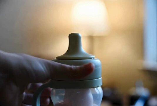 Close up hand gently rotating feeding bottle with handles reveals liquid inside in soft light indoor setting at slow pace baby care concept
