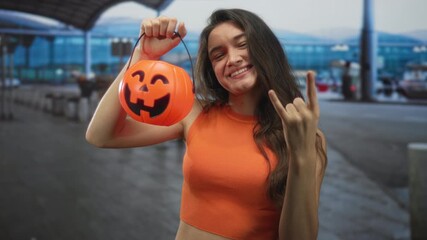 Young hispanic woman holds orange pumpkin bucket and makes rock horns at busy airport terminal; festive energy. - Powered by Adobe