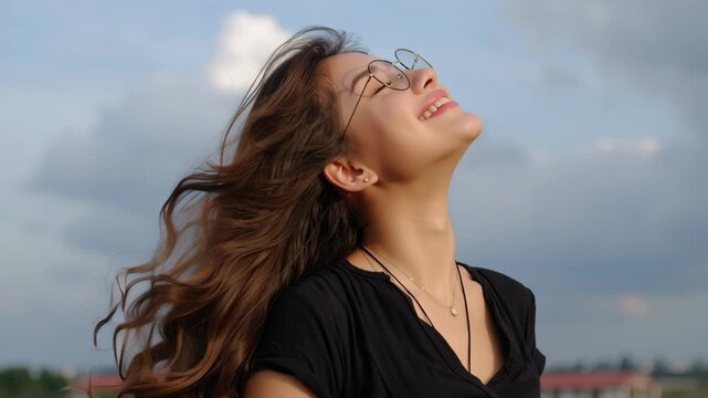 Young woman smiling with flowing hair wearing glasses and necklace outdoors sunset, wind hair with sunlit face and relaxed joyful expression relaxed