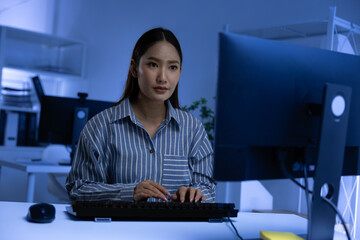 Asian young woman working at computer at night in modern office focused expression illuminated by blue light