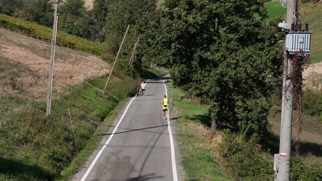 Man strolling with his great dane dog along a winding country road in terraced vineyards on a sunny day, enjoying outdoor companionship and wellness while a jogger runs oout of the scene