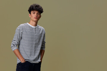 young man wearing striped shirt with hands in pockets stands confidently against muted olive...