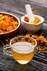 A calendula herbal infusion in a clear glass cup surrounded by dried flowers and natural utensils.