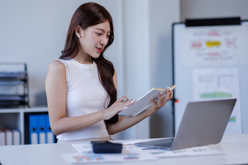 Business woman Asian accountant smiles while analyzing financial data on her laptop and using a calculator in a bright office setting
