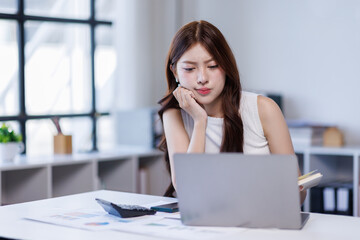 Business woman Asian accountant smiles while analyzing financial data on her laptop and using a calculator in a bright office setting