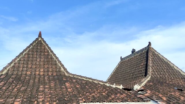 The roof of a traditional Javanese Limasan house with two brown tile roofs with triangular peaks against a blue sky and thin clouds. 