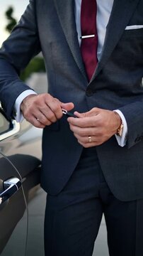Man in suit holding car keys while standing beside luxury vehicle in front of modern building, vertical footage