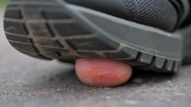 A close-up shot of a dark shoe sole pressing down on a small, reddish candy on a concrete surface, about to crush it.