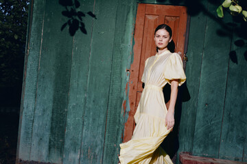 Fashion model wearing a flowing yellow dress stands beside a green wall outdoors, creating a...