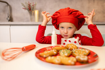 Young child wearing a red chef hat prepares to decorate festive cookies in a modern kitchen during the holiday season