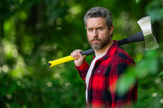Serious lumberjack holding axe and looking away outdoors. Lumberjack worker. A lumberjack with an ax cutting trees in the forest. Man woodcutter outdoor portrait. Concept of a professional logging.