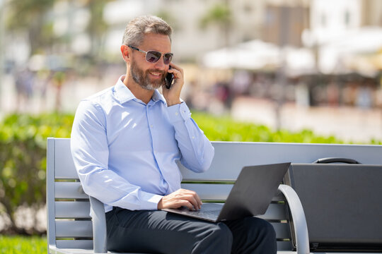 Confident businessman sitting on a bench with a laptop and phone in park. Successful businessman. Mature businessman enjoying work break. Businessman taking a break on a park bench. Summer business.