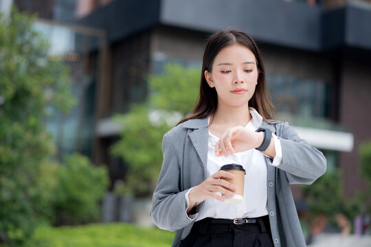 Young asian businesswoman checking time on smartwatch while waiting on busy city street with stress and worried, professional business woman managing schedule and appointment in business district. - Powered by Adobe