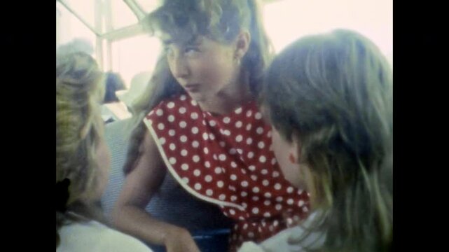 Gossip girl tells friends news. Young female in red polka dot dress enthusiastically talking with interlocutor while traveling on bus. School trip. Old film. Archival retro 1960s. Vintage archive.