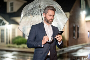 A businessman talks on his phone under an umbrella on a rainy night. Manager makes a business call in the rain. Man in suit speaking on a mobile phone in night street. Business man checks his schedule
