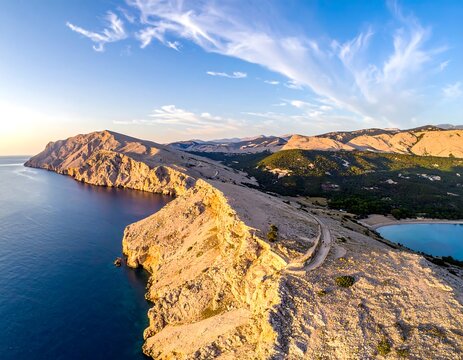 Aerial view of a rugged coastline meeting a deep blue ocean under a bright, partly cloudy sky. The terrain is rocky and sunlit