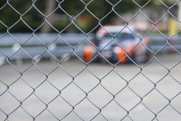 Selective focus on metal chain link fence with wire mesh pattern. blurry orange car behind barrier creates feeling of separation, confinement, and urban boundary