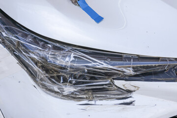 Close up of crashed white car bumper showing significant damage from vehicle accident. makeshift, temporary repair with clear adhesive tape holds broken, scratched plastic together