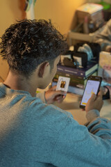 A boy is checking gold price charts on a smartphone while holding a gold bar package at a desk with books and a clock in the background, shot in portrait view from behind