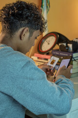 A boy is checking gold price charts on a smartphone while holding a gold bar package at a desk with books and a clock in the background, shot in portrait view from beside