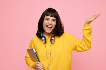A bright portrait of a joyful young woman in a yellow sweater, wearing headphones, holding a tablet and clipboard against a pink studio backdrop, radiating energy and creative zest.