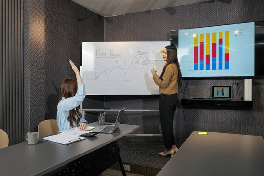Meet business women presenting project work to busy team, people collaborate during meeting, one woman explains data on whiteboard, another raises hand for input, sharing ideas in office