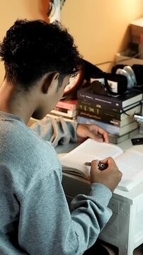 A young man studies at his desk, reading a book with full focus in a cozy indoor setting surrounded by stacks of books, in portrait