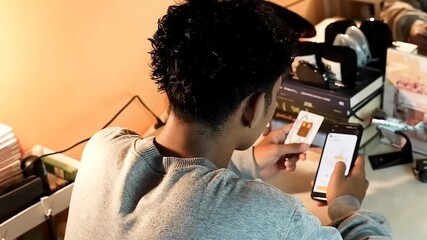 A boy is checking gold price charts on a smartphone while holding a gold bar package at a desk with books and a clock in the background, in landscape view from behind - Powered by Adobe