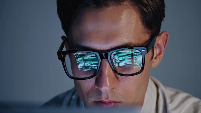In a dimly lit room, a skilled programmer intently reviews lines of code on their computer screen. The reflections in their glasses show complex data, highlighting their concentration and dedication