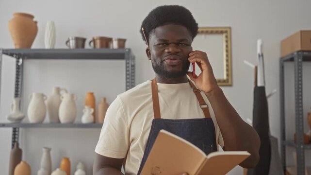 Man in denim apron talking on smartphone while holding notebook with pencil tucked behind ear in studio; focused craft.