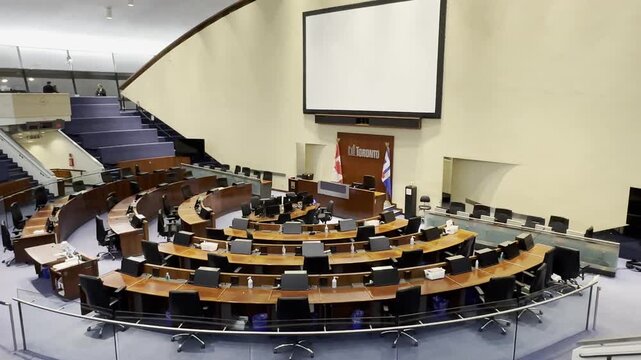 An elevated view from the public gallery looking down at the unoccupied Toronto City Council Chamber. The presentation screen and mayor's desk are visible