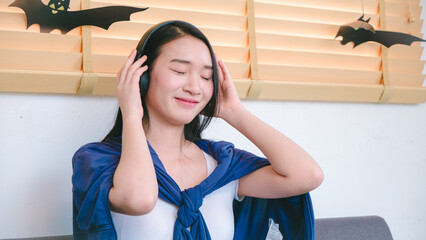 Young woman enjoying music with headphones, smiling happily in a cozy room decorated for Halloween with bats on the walls and a relaxing atmosphere