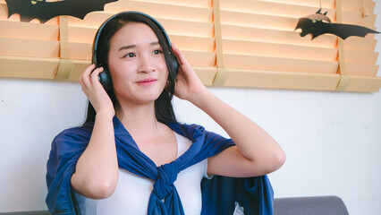 Young woman in blue cape listening to music with headphones in cozy room decorated for Halloween with paper bats on window and warm atmosphere