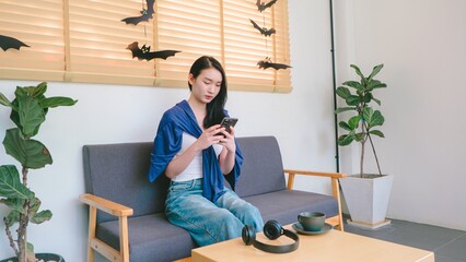 Young woman sitting on sofa in cozy room with greenery, checking smartphone amidst decor of bats and autumn ambiance