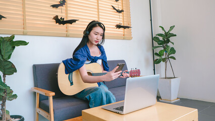 Young woman sitting on a couch playing guitar and using a smartphone while wearing headphones in a modern indoor setting with creative decor