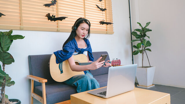 Young woman sitting on a couch playing guitar and using a smartphone while wearing headphones in a modern indoor setting with creative decor