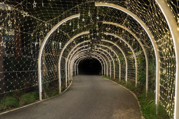 A curved tunnel lined with warm lights glows softly in the dark, and the illuminated arches guide the walkway through a quiet night scene.