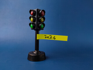 This image features a miniature traffic light toy with a yellow sticky note attached to its pole that has the year 2026