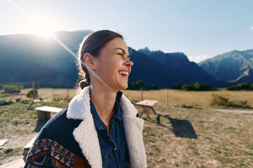 Fototapeta premium Woman smile laughing outdoors in sunlight with mountains backdrop, wearing warm jacket and denim shirt, candid portrait enjoying nature in a meadow by picnic tables and open field.