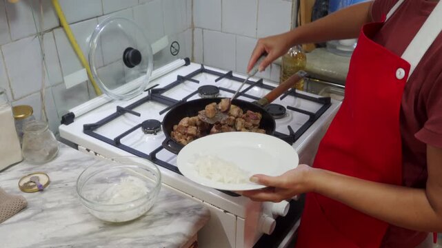 A white plate containing fried pork and onions is held by a cook. She uses a metal spatula to serve the hot meat from the skillet for a delicious dinner.