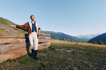 Fototapeta premium Woman in jacket and boots standing by a wooden fence in countryside meadow with mountains in background, embracing nature and wide landscape in a sunlit outdoor relaxed scene for lifestyle.