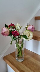 A tall, clear glass vase containing a bouquet of wilted roses in pink, red, and white hues is positioned on light wooden stairs. The roses show signs of wilting, with some petals curled and drooping