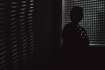 Silhouette of a woman in a dim room touching her forehead near closed blinds, dramatic patterned light symbolizing emotional tension, solitude and deep personal reflection.