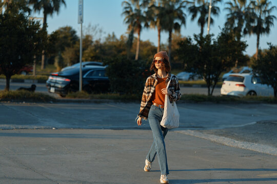 woman walking tote sunglasses golden hour candid casual parking lot. Young woman strolls at sunset with authenticity and candid lifestyle vibe, golden hour glow, mindful living and emotional - Powered by Adobe