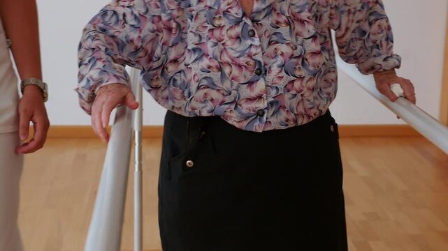 Elderly woman practices walking on parallel bars under the supervision of a physiotherapist. Leg rehabilitation, balance, and walking safety at an active geriatric center.