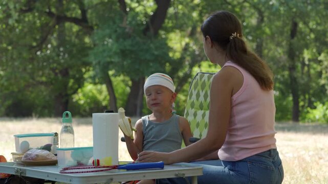 A yellow banana is eaten by the hungry boy sitting in a chair. The caring mother watches him across the aluminum table during their outdoor lunch.